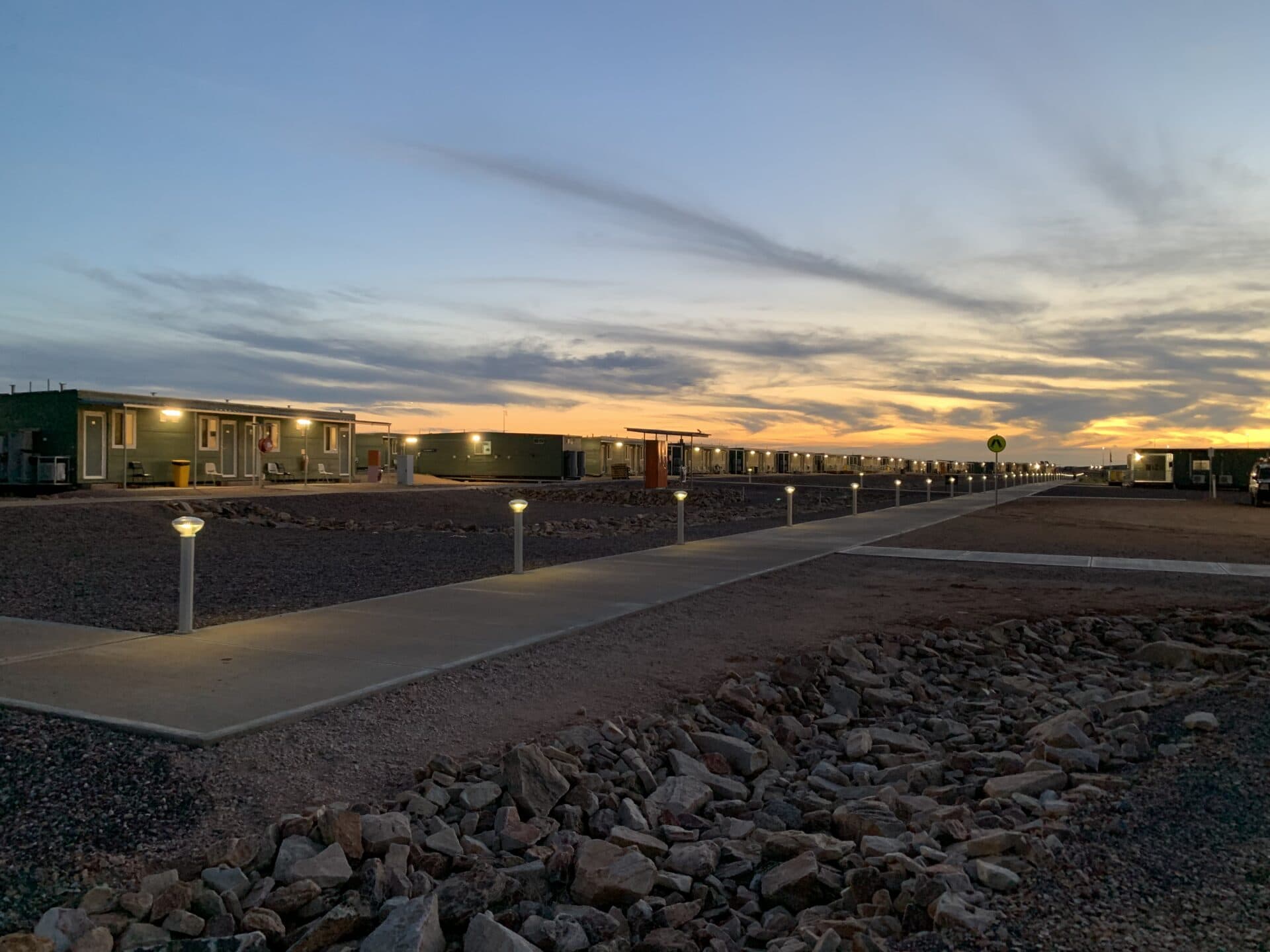 A row of low buildings with bright path lights at sunset, set in a rocky, flat terrain under a cloudy sky.