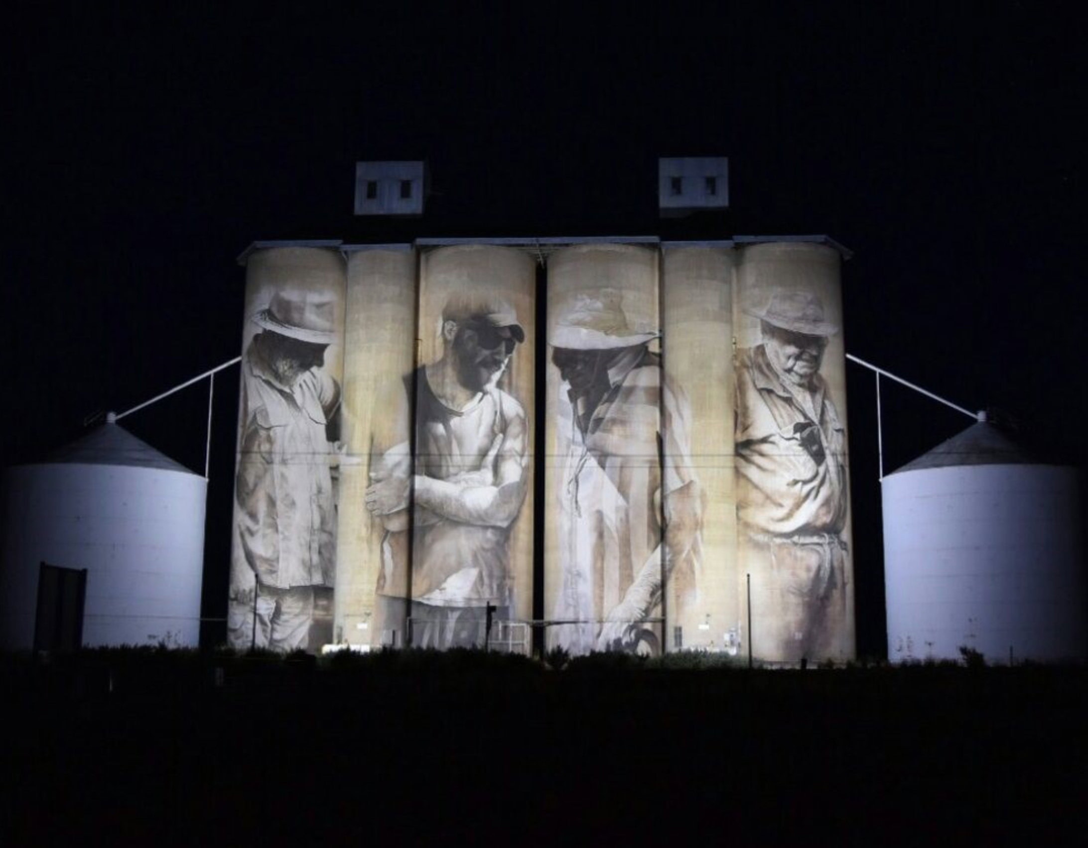 Grain silos at night featuring large murals of four individuals in work attire.