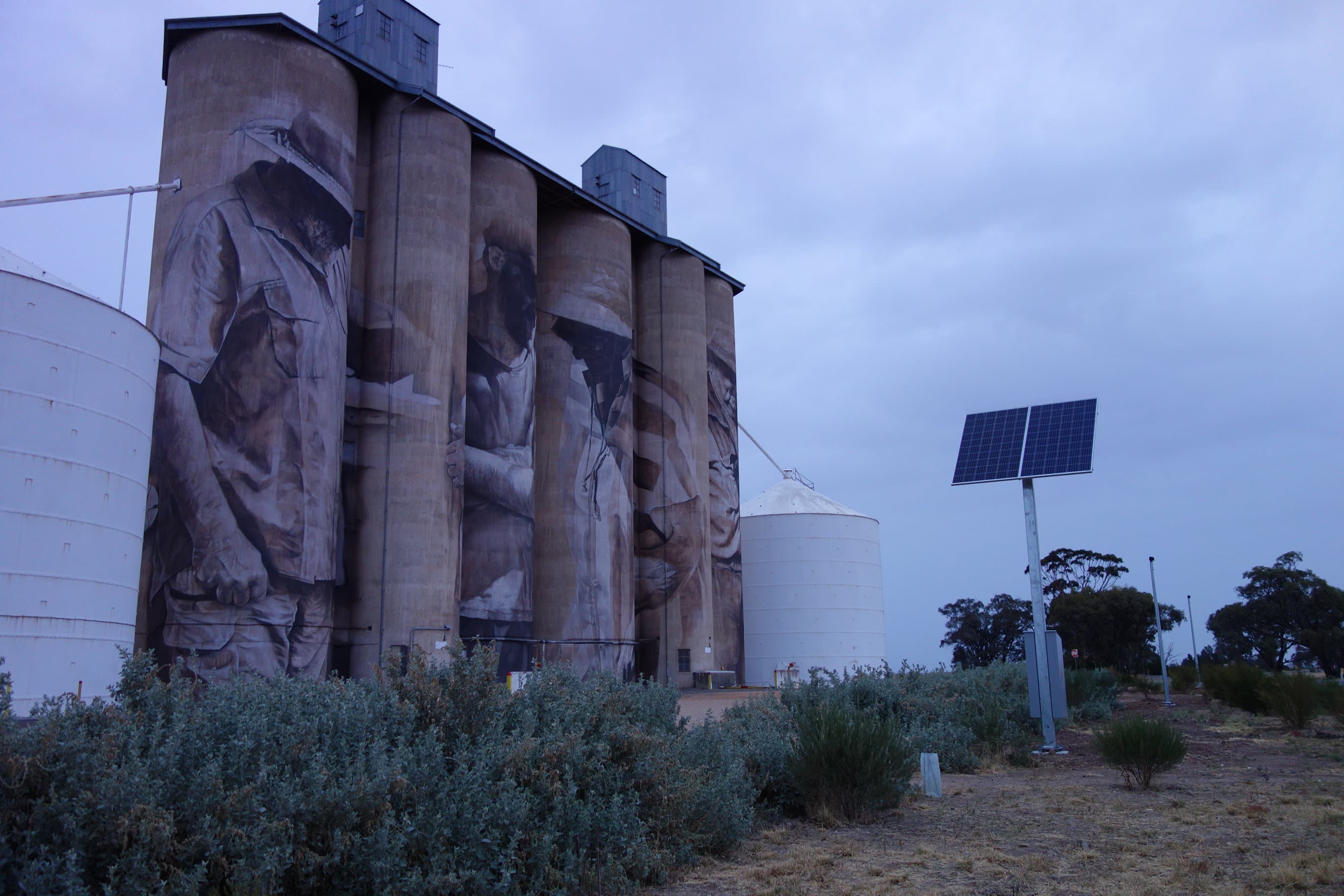 Large silos with artistic murals are surrounded by greenery. A solar panel stands nearby under a cloudy sky.