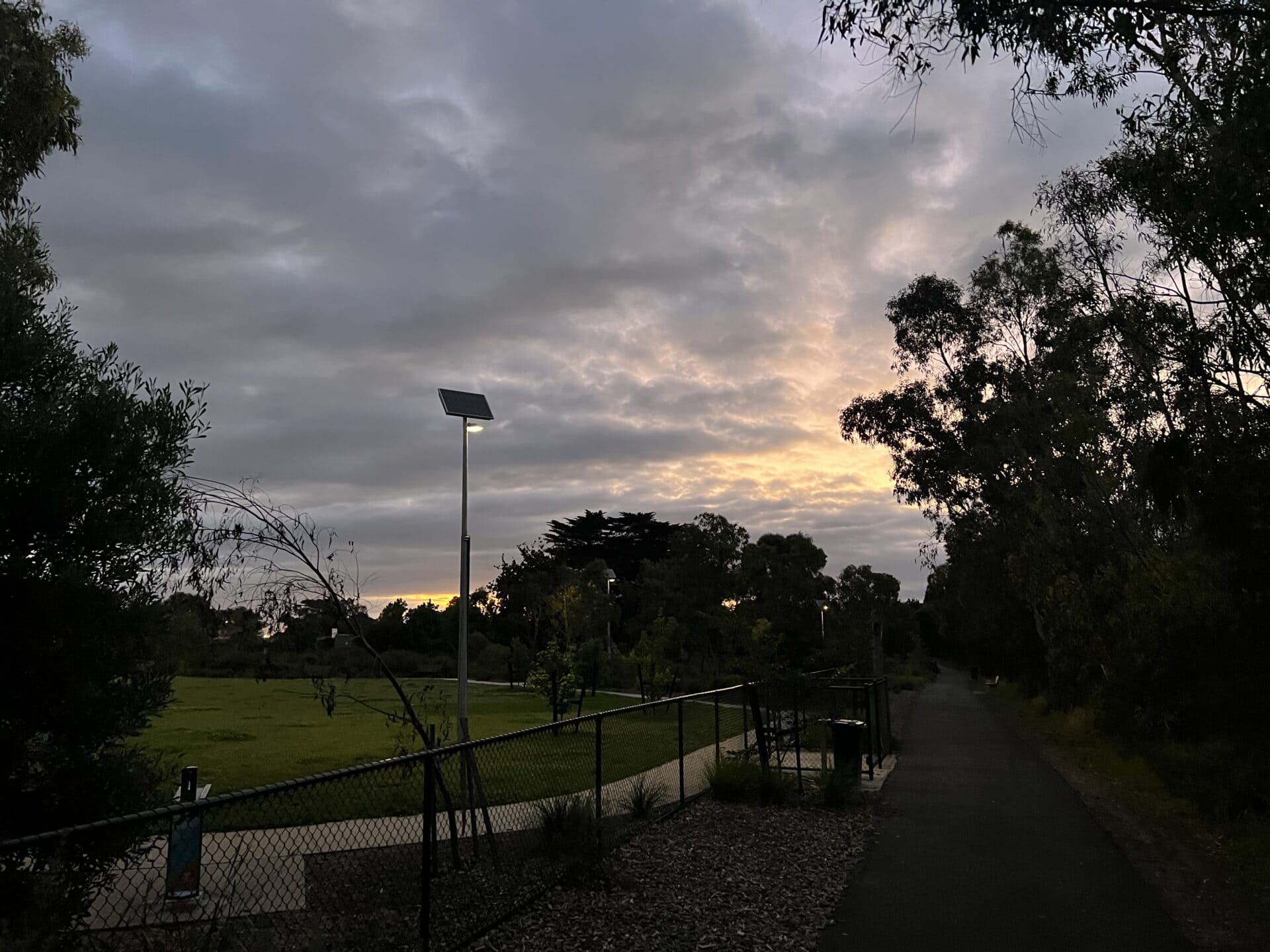 A paved path with a fence and solar light pole at dusk, surrounded by trees and grass, under a cloudy sky.