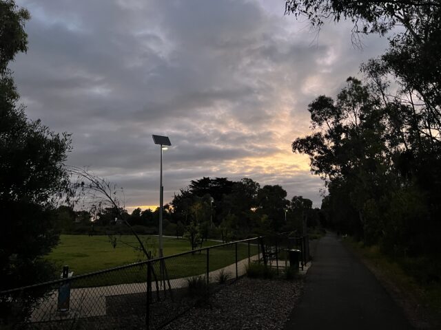 A paved path with a fence and solar light pole at dusk, surrounded by trees and grass, under a cloudy sky.