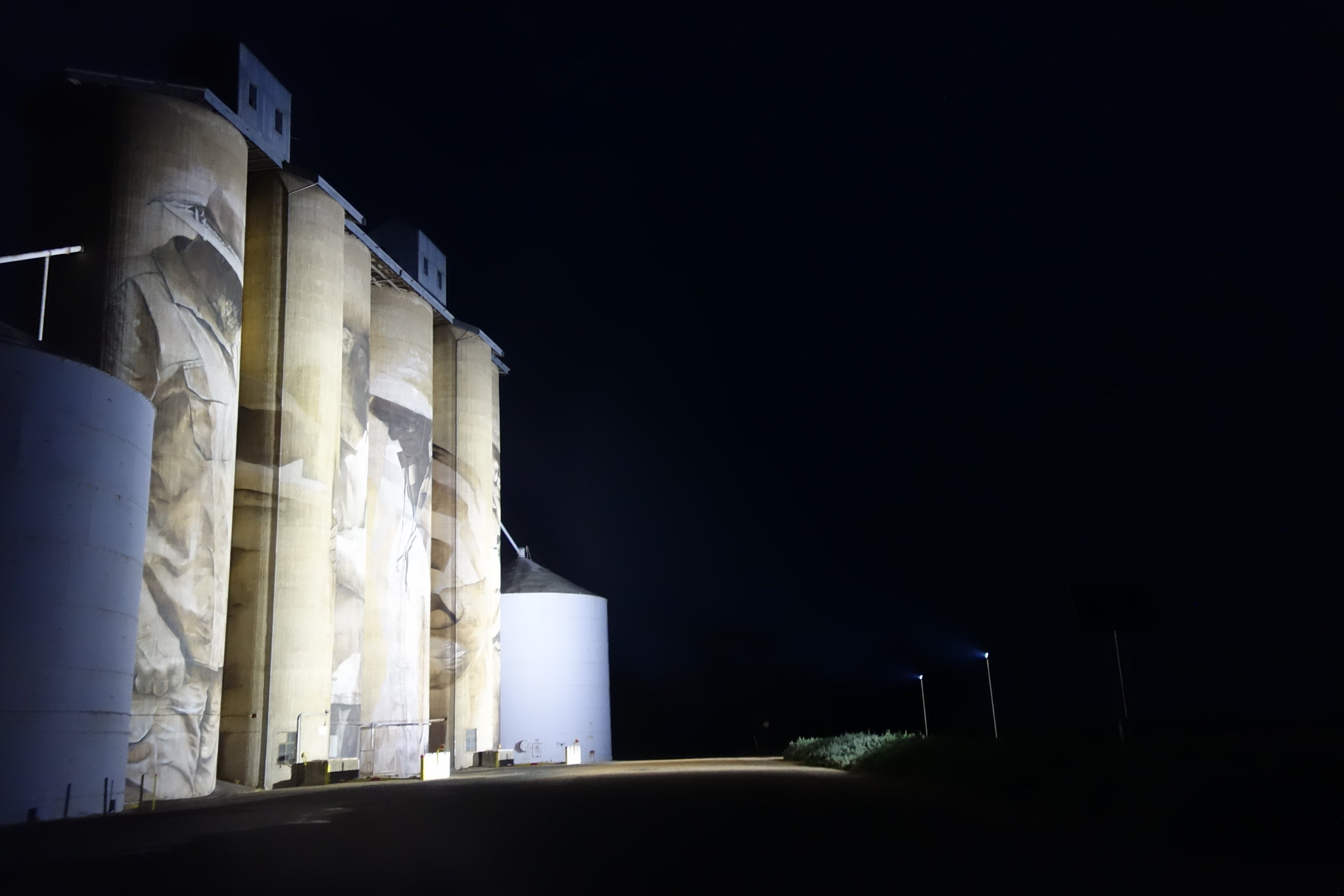 Tall grain silos are illuminated at night, with some faint artwork visible on the structures. Dark sky and ground add contrast, highlighting the silos' architectural features.