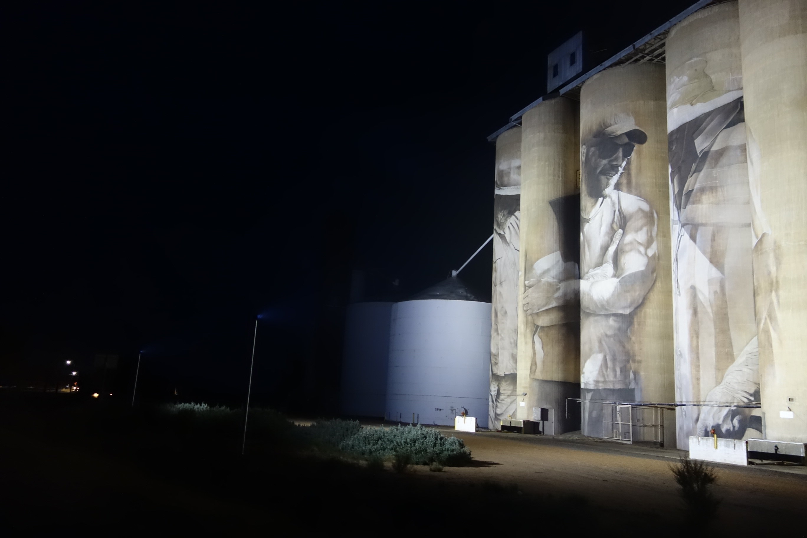 Night view of large grain silos with murals depicting people in work attire. A smaller silo is visible beside them.